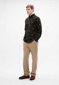 Mørkegrøn kordfløjlskjorte med fuglemønster, beige bukser og brune ruskindsko. Outfittet har en knap-ned krave og en tætsiddende Silhuet.