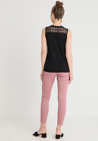 Kvinna stående vänd bort, iklädd svart ärmlös topp med spetsdetalj och rosa smal passform byxor med svarta öppna klackskor.