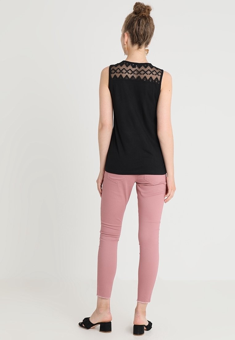 Kvinna stående vänd bort, iklädd svart ärmlös topp med spetsdetalj och rosa smal passform byxor med svarta öppna klackskor.
