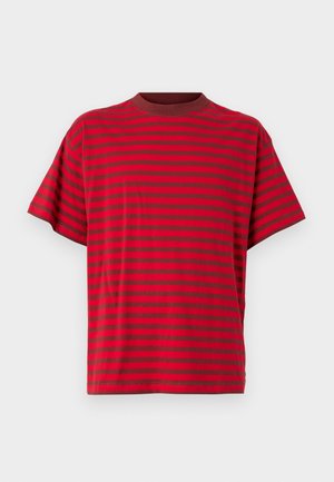 Personne portant une chemise à manches courtes rayée rouge et rouge foncé, un short noir au niveau du genou, des chaussettes blanches et des baskets noires, debout devant un fond blanc.
