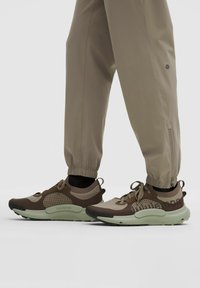 Sportschoenen in een bruin en beige kleurschema, met een bovenwerk van gaas en stof, en een groene rubberen zool; gecombineerd met lichtbeige joggingbroeken.