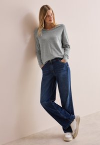 Graues Langarmshirt kombiniert mit locker sitzenden dunkelblauen Denim-Jeans und weißen Sneakers; das Model steht mit Händen in den Taschen vor einem neutralen Hintergrund.