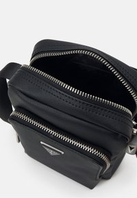 Bolso cruzado negro con dos compartimentos con cremallera, hecho de material texturizado. Presenta herrajes plateados y un acento de logo en la parte frontal.