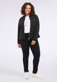 Femeie cu păr creț, îmbrăcată cu o jachetă din denim neagră, o cămașă albă cu grafică, blugi negri și teniși albi, stând în fața unui fundal simplu.