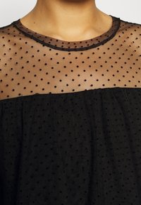 Svart genomskinlig topp med ett prickigt mönster. Har en rundad halsringning med fransiga kanter och textuerad svart tyg under det genomskinliga lagret.