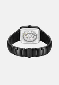 Quadratische schwarze Edelstahl-Uhr mit transparentem Gehäuseboden, wasserdicht bis 5 ATM und einem schwarzen Gliederarmband. Auffällige Markengravur sichtbar.