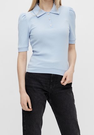 Nainen, jolla on päällään vaaleansininen ribbitekstiilinen lyhythihainen paita, jossa on kaulus ja kolme koristeellista nappia, yhdistettynä korkeavyötäröisiin mustiin farkkuihin.