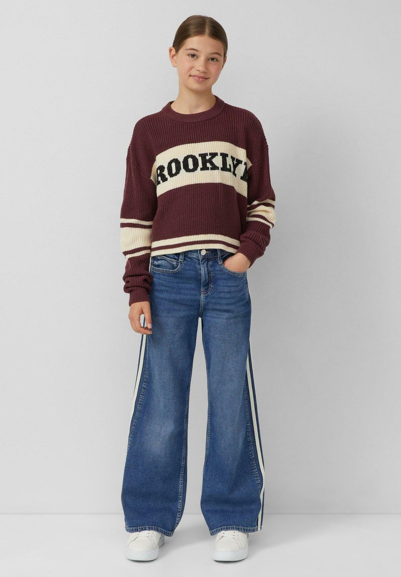 Maglione a maglia color bordeaux con testo "BROOKLYN", abbinato a jeans a gamba larga blu con strisce laterali bianche e sneakers bianche.