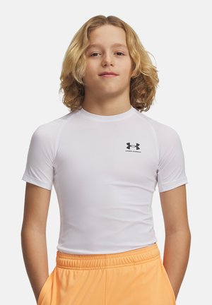 Jongen met schouderlang blond haar, die een wit Under Armour compressieshirt en oranje korte broek draagt, staat met zijn handen in zijn zakken.