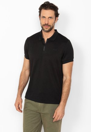 Homme à la barbe portant un polo noir à manches courtes avec une fermeture éclair et un pantalon vert olive, debout devant un fond blanc.