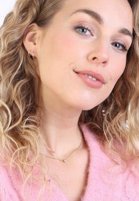 Collier en or avec un petit pendentif, cheveux blonds bouclés, portant des boucles d'oreilles en or et un pull rose doux avec un motif de tricot texturé.