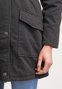 Zbliżenie ręki osoby opierającej się przy dolnej kieszeni klapy czarnej zapinanej kurtki na niebieskich dżinsach.