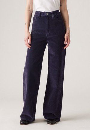 Szerokie spodnie z wysokim stanem z ciemnofioletowego sztruksu, noszone z czarnymi butami i białą, włożoną do środka, bezrękawnikową bluzką.