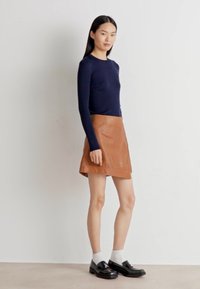 Top a maniche lunghe in maglia blu navy abbinato a una gonna avvolgente in pelle beige, calze bianche alla caviglia e scarpe nere, presentato da una modella su uno sfondo chiaro.