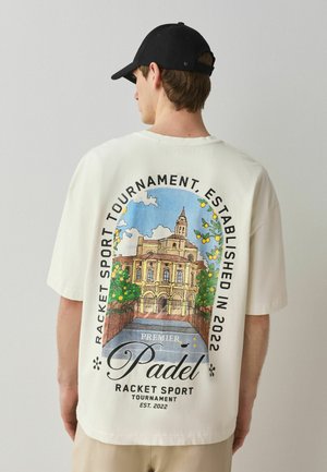 Young man wearing a white oversized t-shirt with a colorful padel racket sport tournament graphic and text on the back.