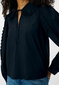 Blusa nera con colletto scalloped, dettaglio a cuore, e maniche a sbuffo con ritagli scalloped. Realizzata in tessuto liscio.