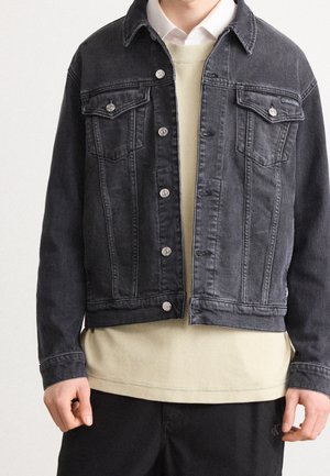 Personne portant une veste en jean noire par-dessus un pull beige et une chemise blanche à col, montrée des épaules aux hanches sur un fond uni.