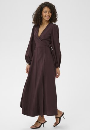Kobieta o kręconych włosach, ubrana w długą sukienkę kopertową w kolorze głębokiego śliwkowego oraz czarne sandały na obcasie, uśmiecha się i spogląda w bok.