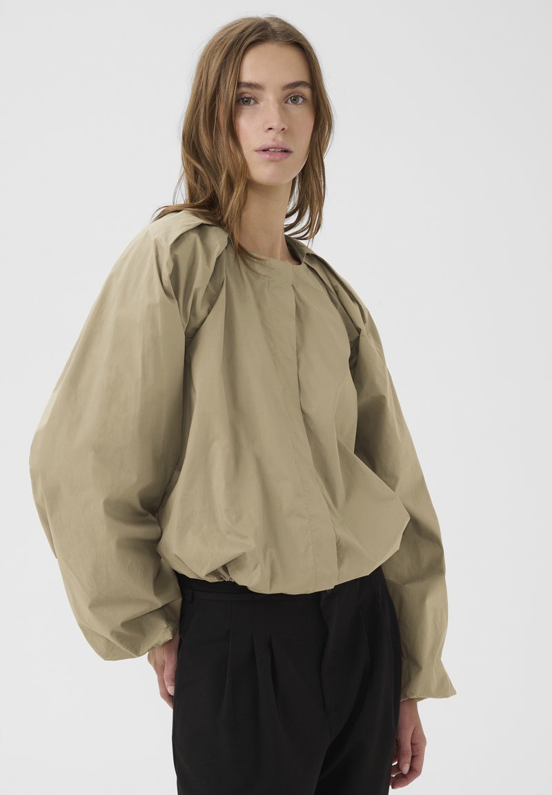 Jeune personne portant un chemisier beige à manches bouffantes surdimensionnées et un pantalon plissé noir, debout devant un fond clair uni.