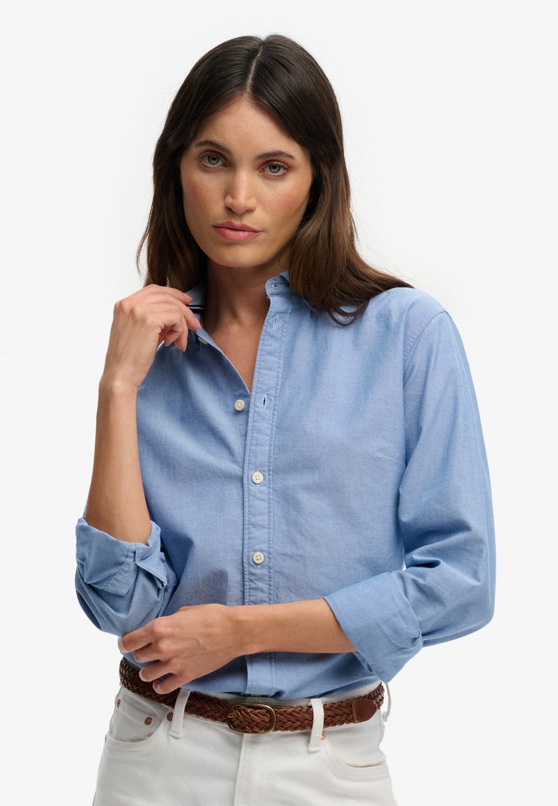 Femme aux longs cheveux bruns, vêtue d'une chemise à boutons bleu clair, d'un pantalon blanc et d'une ceinture tressée marron, ajustant son col.