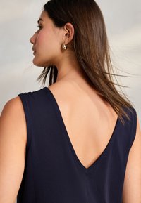 Marinblå ärmlös topp med djup V-formad ryggdesign, med mjukt tyg och ren sömnad. Hår delvis synligt, med texturerade slingor.