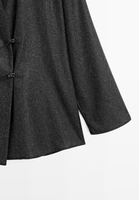 Mørkegrå uldfrakke med en broget tekstur, der har brede ærmer og sidelæg, fastgjort med sorte spænder.