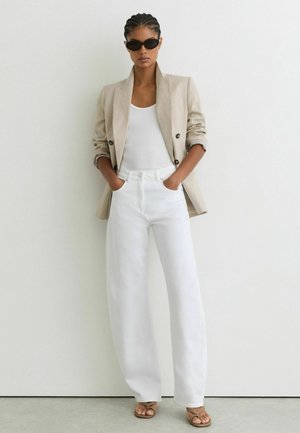 Mujer con blazer beige, camiseta blanca, vaqueros blancos de pernera ancha de talle alto, sandalias marrones y gafas de sol negras, de pie frente a una pared blanca.