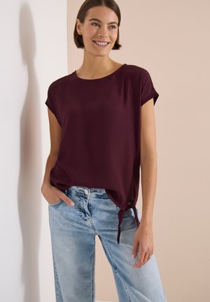 Frau lächelt, trägt eine kurzärmelige dunkelviolette Bluse mit Seitenband und hellblaue Jeans, steht vor einem neutralen Hintergrund.
