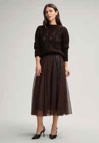 Brązowy sweter dziewiarski o luźnym kroju i teksturowanym wzorze, zestawiony z długą, przezroczystą brązową spódnicą z tiulu z czarnym pasem. Czarne obcasy.