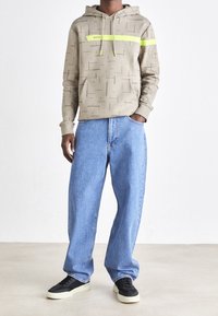 Beige hoodie med geometriska mönster, neongröna detaljer och en framficka, ihop med ljusblå baggy jeans och svarta sneakers.