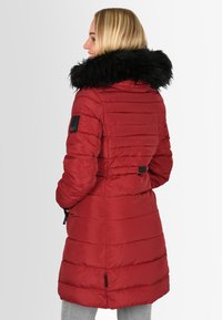 Red padded coat with black fur collar, vertical stitching, adjustable waist, and logo patch on the sleeve. Smooth texture and knee-length.