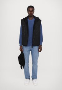 Black sleeveless vest with a textured surface over a blue sweater; light blue jeans and white sneakers. Holds a black bag. White background.