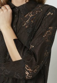 Sort bluse i blonde med krave, knaplukning og tekstureret stof, med gennemsigtig blonedetaljering på ærmerne.