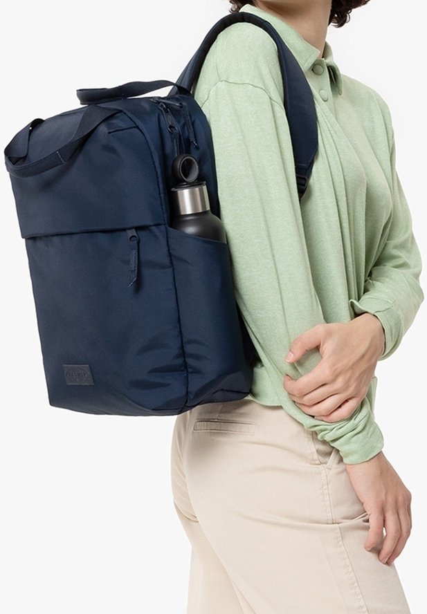 Personne portant une chemise à manches longues vert clair et un pantalon beige porte un sac à dos bleu marine avec une poche latérale contenant une bouteille d'eau noire et argentée.