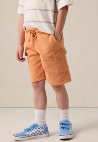 Shorts en polaire orange avec une taille élastique et une poche latérale. Associés à des sneakers à carreaux bleus et blancs et des chaussettes blanches de hauteur intermédiaire.