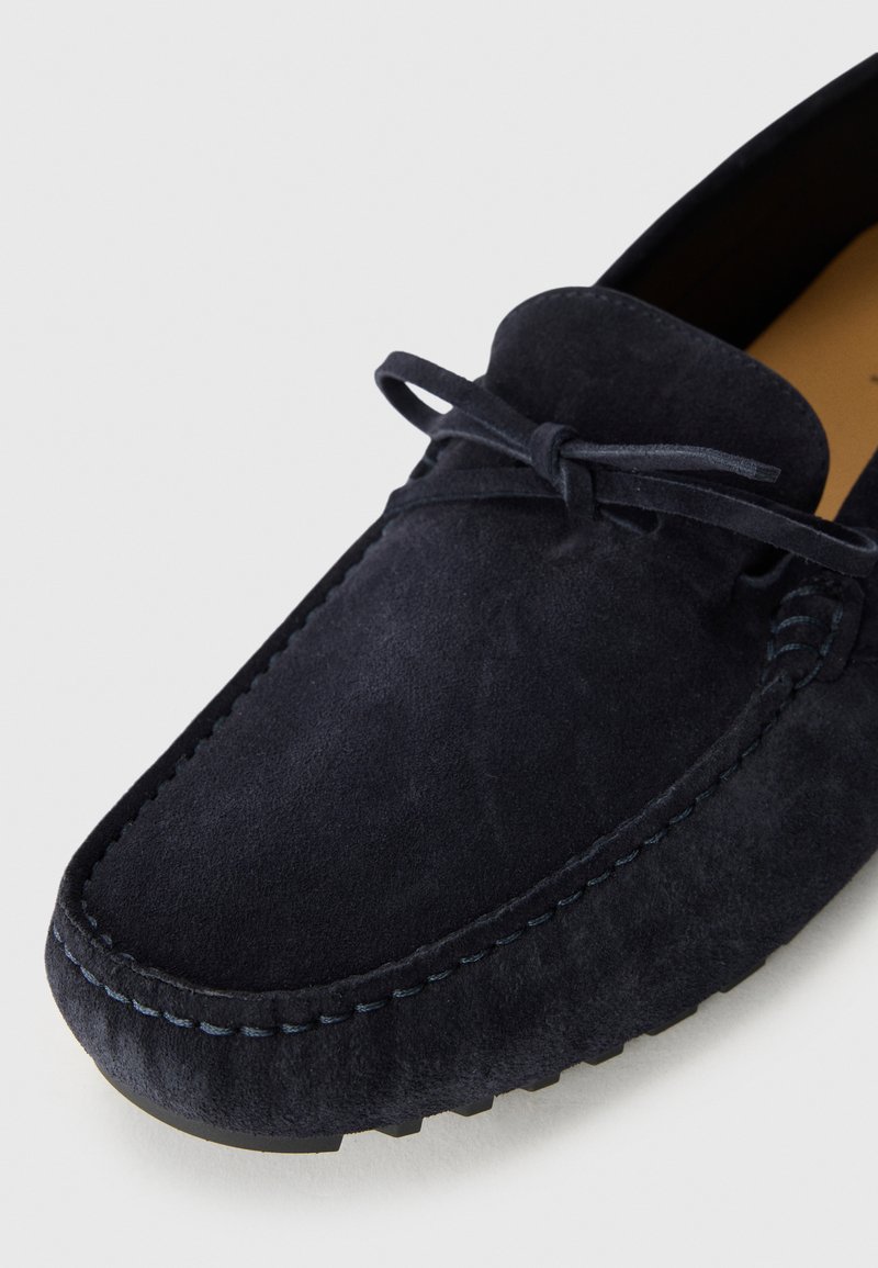 Navy blue suede moccasin shoe with a leather interior, featuring a front bow tie detail and contrasting stitch accents along the sole.
