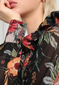 Bloemige blouse van zwart doorschijnend materiaal, met rode en gele bloemen, groene bladeren en een ruffle-kraagdetail op de mouwen.