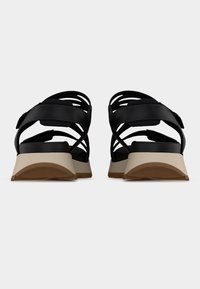 Paire de sandales noires à brides avec talons compensés beige et semelles en caoutchouc, vues de dos sur un fond blanc uni.