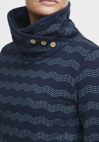 Mørkeblå topp med høy krage og tre treknapper. Har sikksakk hvite mønstre. Laget av teksturert stoff, kroppsnær design.
