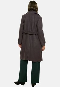 Cappotto viola scuro al ginocchio con collo e cintura in vita. Presenta una texture liscia e una vestibilità su misura, abbinato a pantaloni verde a gamba larga.