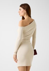 Beige off-the-shoulder dress with long sleeves, form-fitting silhouette, smooth texture, and a side slit, held with a small dark clutch.