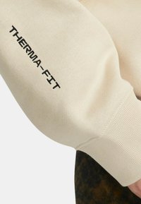 Felpa beige con ricamo "THERMA-FIT" in nero sulla manica. Texture liscia con dettagli a costine sui polsini. Design minimalista.
