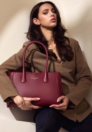 Sac à main en cuir bordeaux avec deux longues poignées, une texture lisse et un logo en relief. Porté avec un blazer marron et un jean foncé.