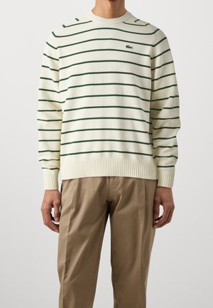 Uomo che indossa un maglione crema con righe orizzontali verdi e pantaloni kaki su uno sfondo chiaro e semplice.