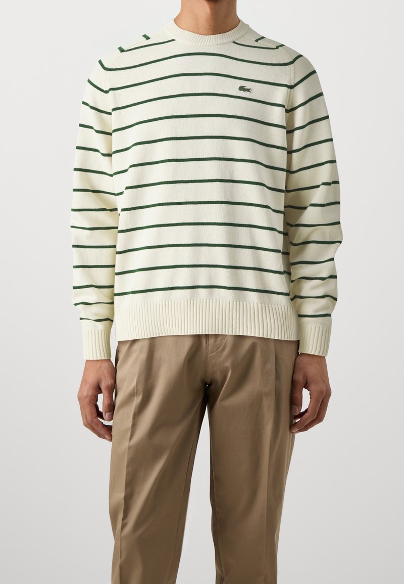Homme portant un pull crème avec des rayures horizontales vertes et un pantalon kaki sur un fond clair uni.