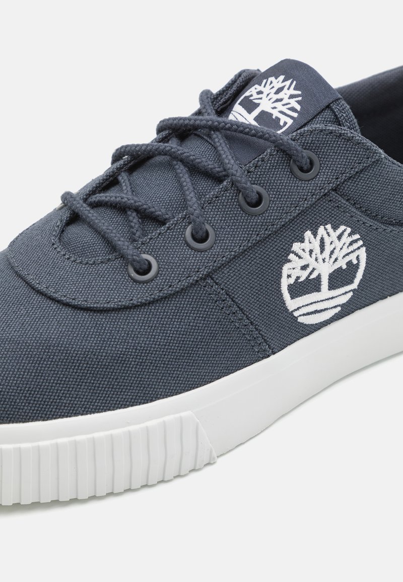 Zapatillas de lona azul oscuro con suela de goma blanca, punta redonda, cordones planos y parche con el diseño de un árbol en el lateral.