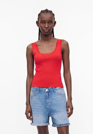 Mujer con el pelo trenzado que lleva una camiseta sin mangas roja con borde ondulado y shorts vaqueros azul claro doblados, de pie frente a un fondo liso.