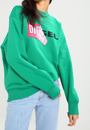 Personne portant un sweat-shirt vert vif avec un logo graphique rouge et noir et un jean bleu clair sur un fond clair uni.