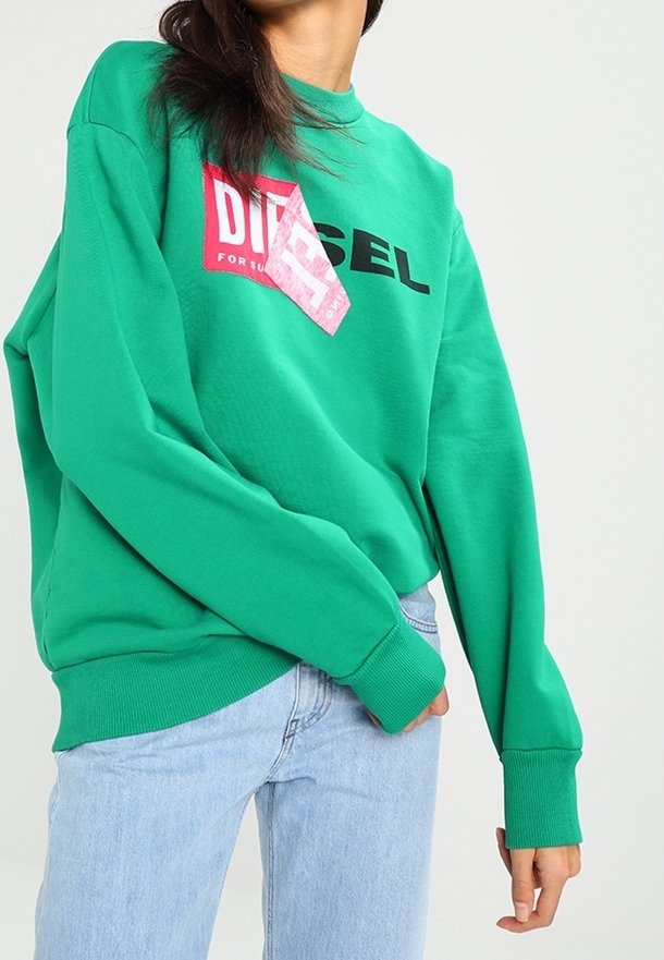 Personne portant un sweat-shirt vert vif avec un logo graphique rouge et noir et un jean bleu clair sur un fond clair uni.