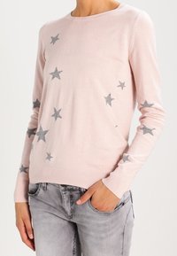 Pull-over rose clair à manches longues avec des motifs d'étoiles grises dispersées, porté avec un jean gris clair, main reposant dans la poche avant.